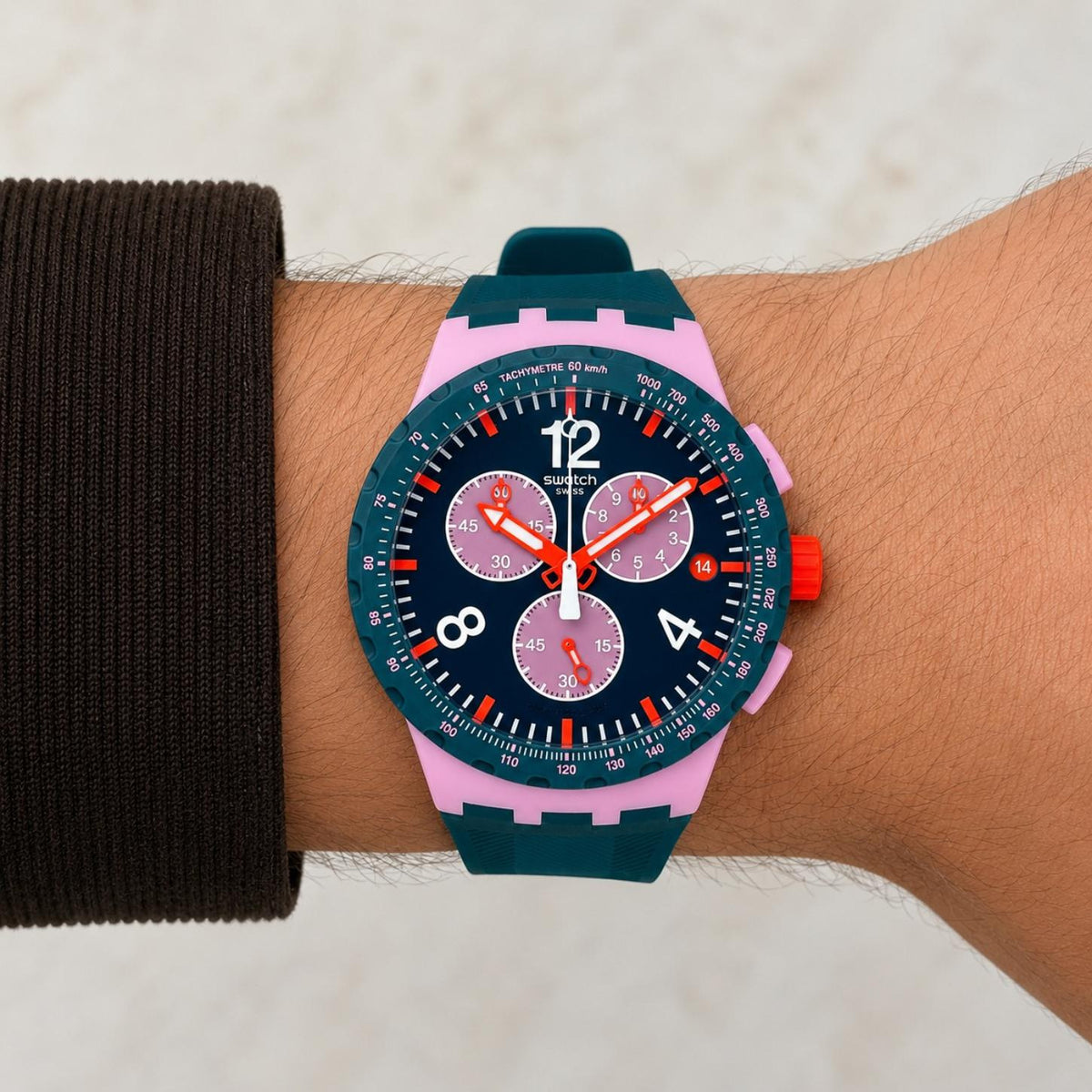Swatch Moment of Rose SUSP400 worn on the wrist with green chronograph dial, three subdials, white numerals, orange hands and accents, pink case, and dark green strap.