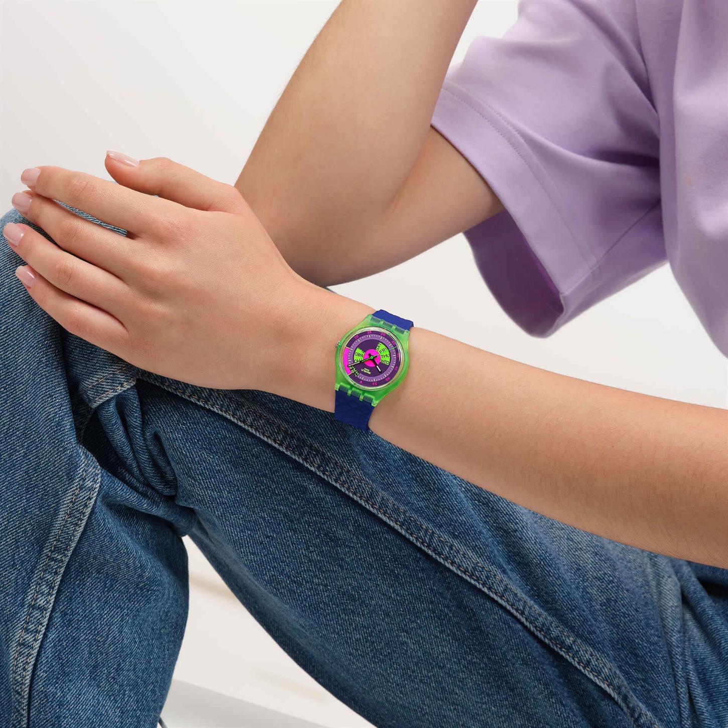 Swatch Neon Skychart SO28G704 watch with a purple dial, translucent green case, colorful day display, and blue strap worn on the wrist.