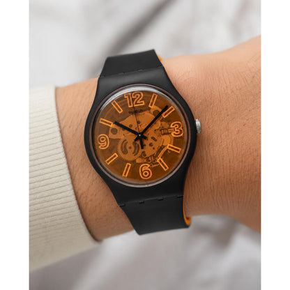 Swatch Orangeboost SUOB164 watch with a transparent orange dial, visible movement, black case, orange numerals, and black strap worn on the wrist.