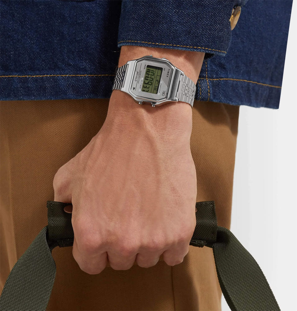 Reloj Timex digital de acero lucido en la muñeca de una persona sosteniendo una bolsa, estilo casual.