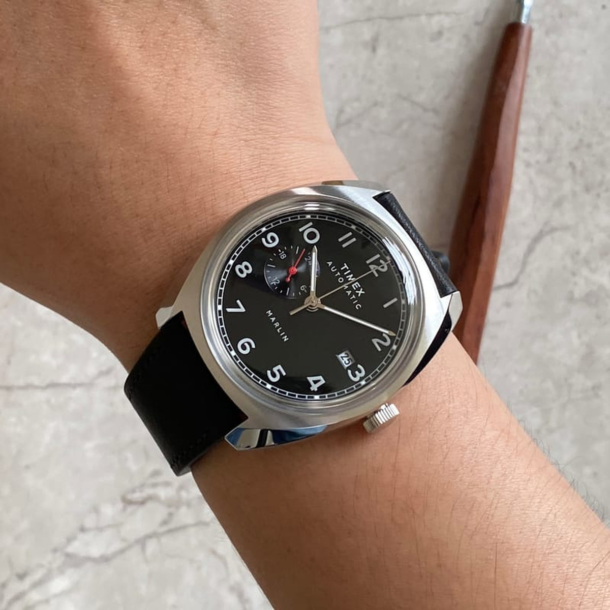 Timex Marlin TW2V62100 worn on the wrist with black dial, silver-tone Arabic numerals and hands, 24-hour subdial with red accent hand, date window, polished cushion-shaped case, and black leather strap.