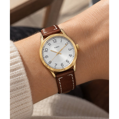 Timex New England TW2R23000 with a silver dial, Arabic numeral hour markers, gold-tone case and hands, and brown leather strap with white contrast stitching worn on the wrist.