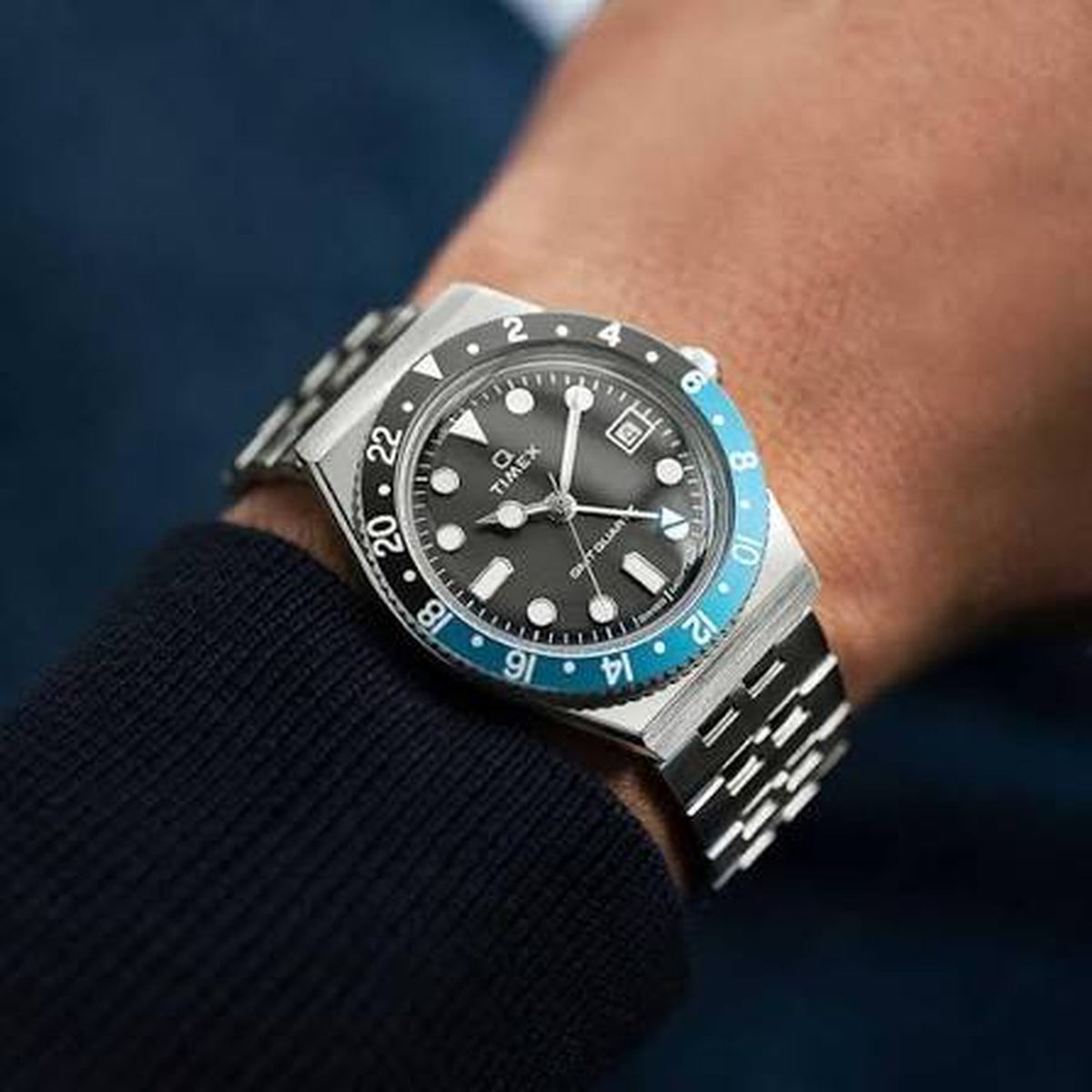 Timex Q Timex GMT TW2V38100 with a black dial, black and blue 24-hour GMT bezel, date window, silver-tone case, and matching bracelet worn on the wrist.