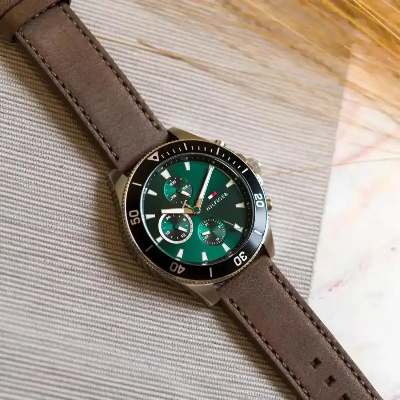 Tommy Hilfiger 1791983 men’s multifunction watch with green dial, black bezel, and brown leather strap displayed on a textured surface.