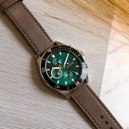 Tommy Hilfiger 1791983 men’s multifunction watch with green dial, black bezel, and brown leather strap displayed on a textured surface.