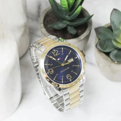 Reloj Tommy Hilfiger con esfera azul y brazalete bicolor apoyado en una superficie clara, rodeado de plantas suculentas decorativas.